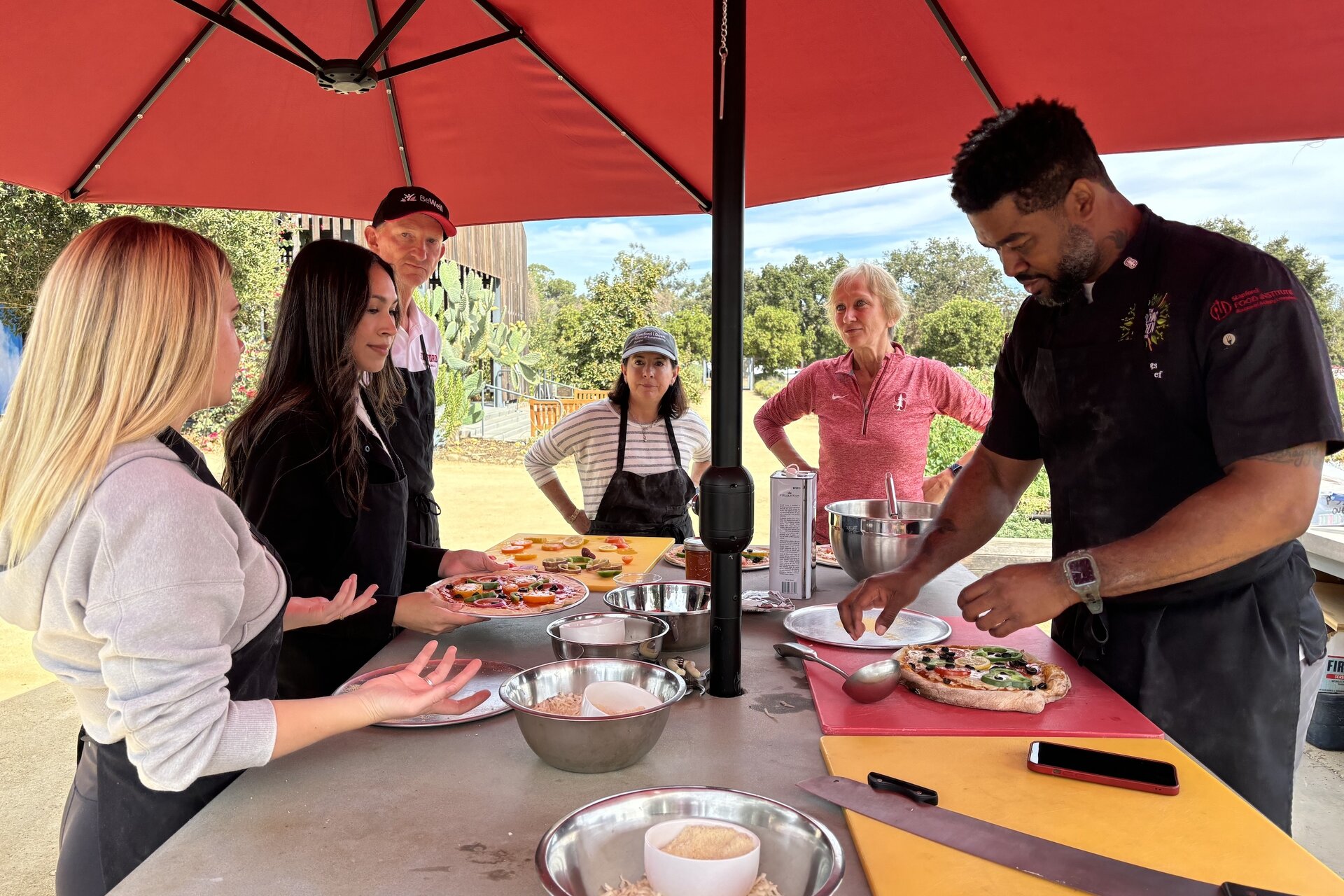 People preparing food around an outdoor table under a sun umbrella
